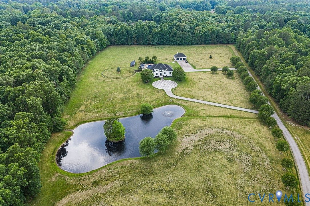 Aerial view of the 20.3-acre Bradley Bridge property showing the white estate, circular driveway, detached workshop, mowed lawns, and a stocked pond ringed by mature Virginia hardwood forest